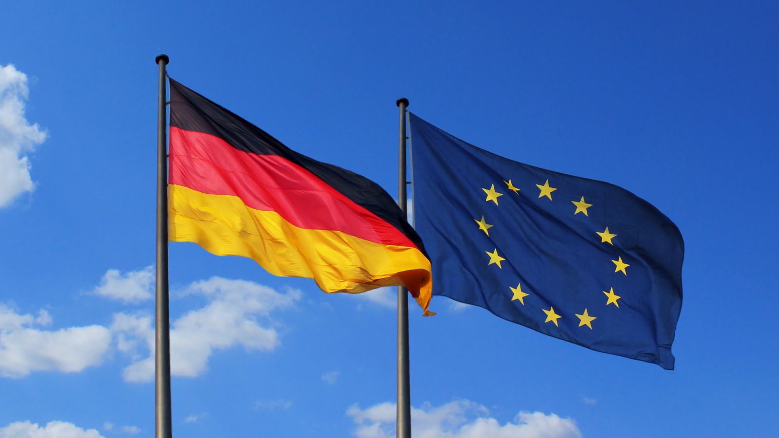 Eine Deutschland- und eine EU-Flagge wehen vor blauem Himmel im Wind.
