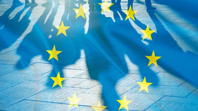 Der Schatten von mehreren Personen fällt auf einen Bürgersteig, davor ist eine transparente EU-Flagge zu sehen.