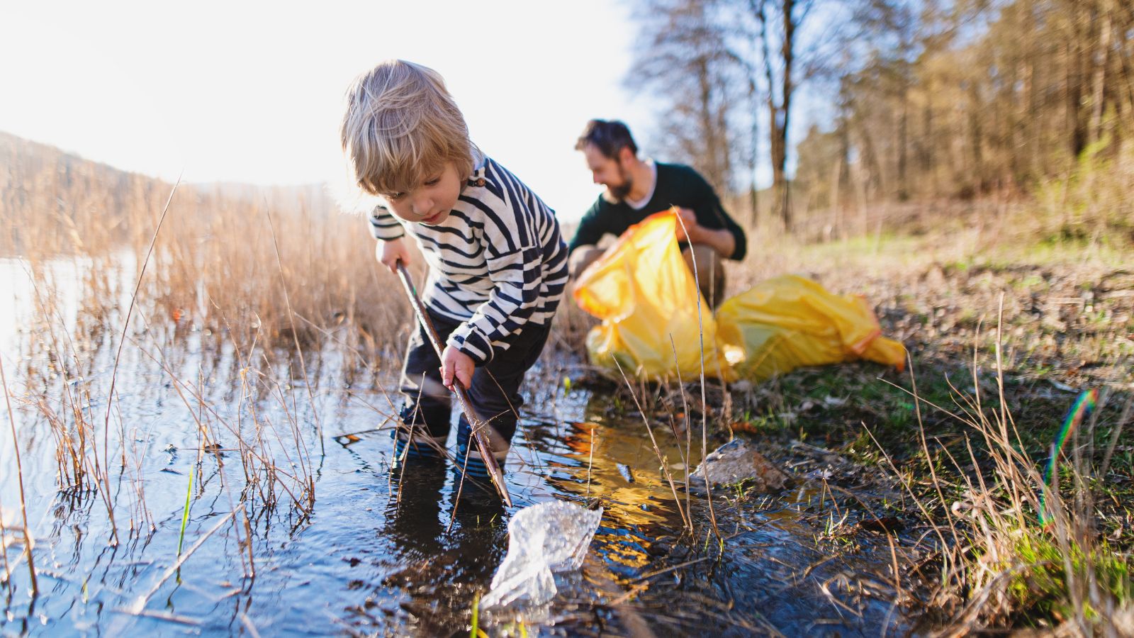 Kind und erwachsener Mann sammeln Müll aus seichtem Gewässer am Uferrand eines Sees, bei strahlender Sonne (
