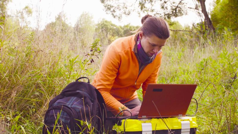 Eine Frau in Trekking-Garderobe hockt im hohen Gras. Neben ihr liegt befindet sich ein Rucksack. Vor ihr steht ein aufgeklappter Laptop auf einer großen Plastikkiste.