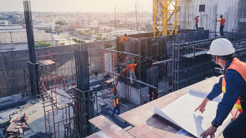 Ein Mann mit Schutzhelm und Warnweste kniet auf dem Dach eines Neubaus. Er betrachtet das geschehen auf der Baustelle während er vor sich einen großen Plan ausbreitet.