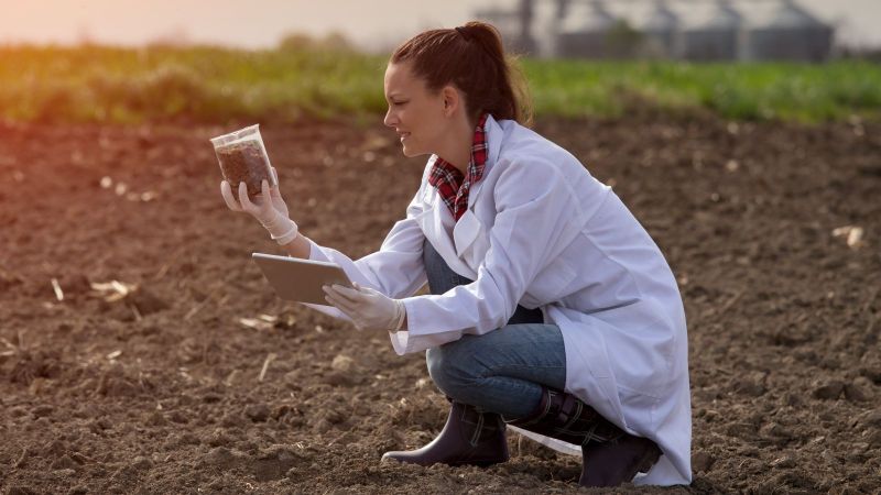 Eine Frau mit weißem Laborkittel kniet auf einem Acker und hält ein Becherglas mit einer Bodenprobe und ein Tablet in den Händen. Im Hintergrund sind eine Wiese und eine Biogasanlage zu sehen.