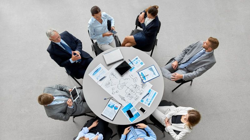 Vogelperspektive auf eine Gruppe von Menschen in formalen Anzügen an einem runden Tisch sitzend. Darauf sind Planungsmaterialien erkennbar. Der Hintergrund ist ein grauer Boden.