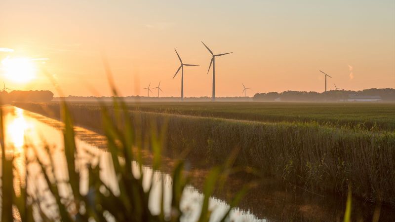 Aufgehende Sonne scheint auf Kanal, welcher durch landwirtschaftliche Felder führt, mit Windkraftanlagen im Hintergrund