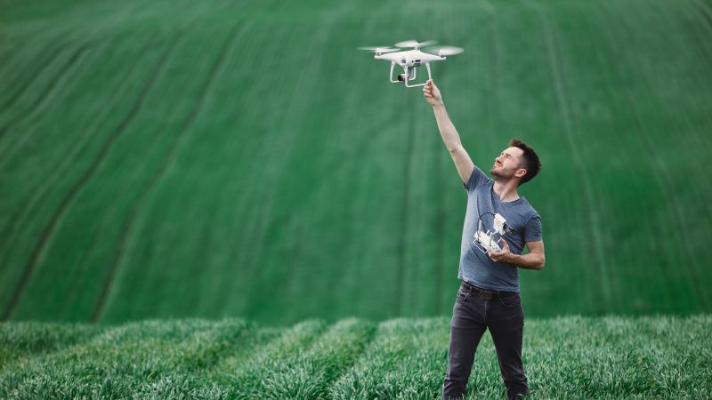Mann steht in weitem Feld mit kniehohem Gras. Er hält eine fliegende Drone in einer Hand hoch und die Fernsteuerung in der anderen.