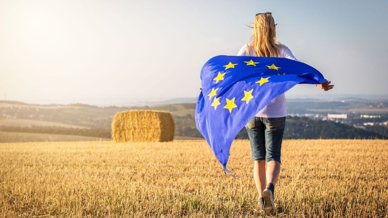 Frau steht auf Stoppelfeld mit dem Rücken zum Betrachtenden und einer Europaflagge hinterm Rücken in beiden Händen tragend.