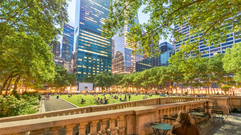 Eine Person sitzt auf einer Außenterrasse vor einem Park mit vielen Menschen, Bäumen an den Seiten und Hochhäusern im Hintergrund