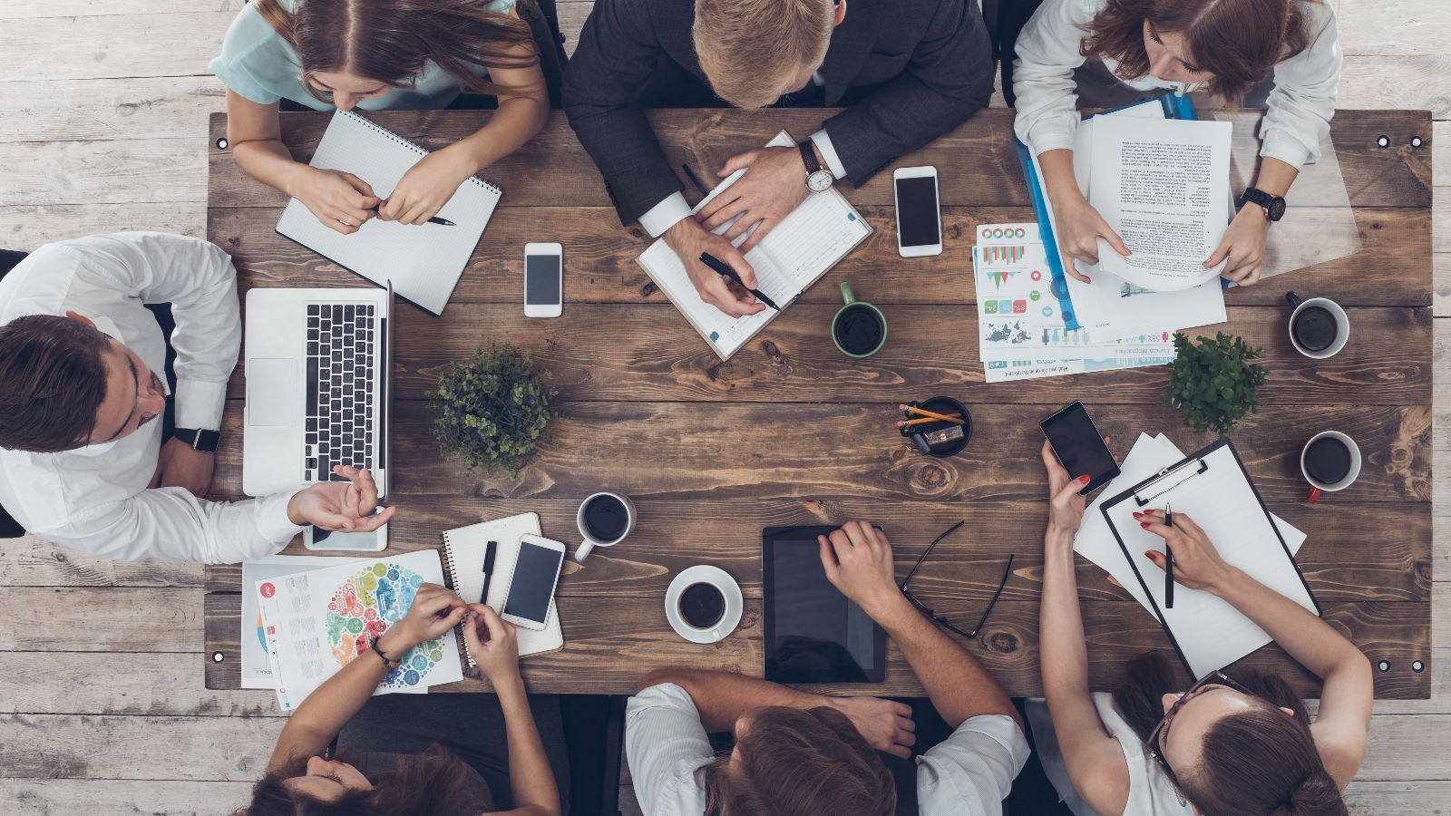 Draufsicht auf einen Tisch mit Unterlagen, Laptops, Tablets, Smartphones und Kaffeetassen. Um den Tisch sitzen sieben Personen und diskutieren.