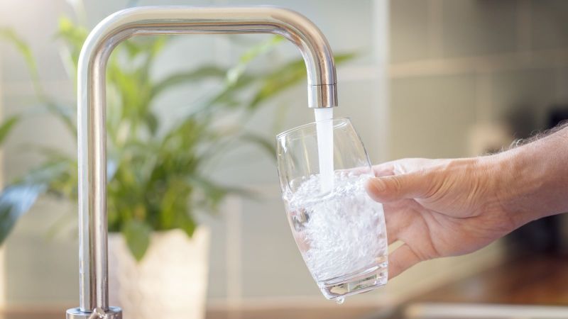 Eine Hand hält ein Glas, das gerade mit Leitungswasser befüllt wird. Im Hintergrund ist unscharf ein heller Raum mit Zimmerpflanze erkennbar. 