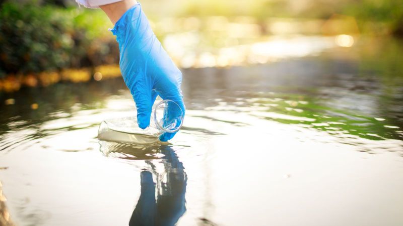 Eine Hand mit blauem Gummihandschuh entnimmt eine Wasserprobe aus einem Gewässer, im Hintergrund ist unscharfes Sonnenlicht