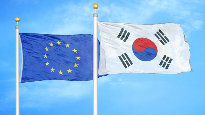 Flaggen der Europäischen Union (links) und Südkoreas (rechts), die nebeneinander an Fahnenmasten vor einem klaren blauen Himmel wehen