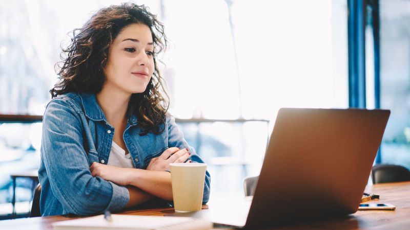 Eine junge Frau sitzt aufmerksam mit Kaffeebecher und Notizblock vor einem aufgeklappten Laptop.