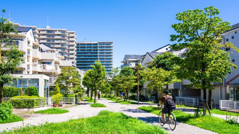 Ein Mann fährt auf dem Fahrrad durch eine begrünte Wohnsiedlung einer Stadt. Im Hintergrund sind hohe Gebäude zu sehen.