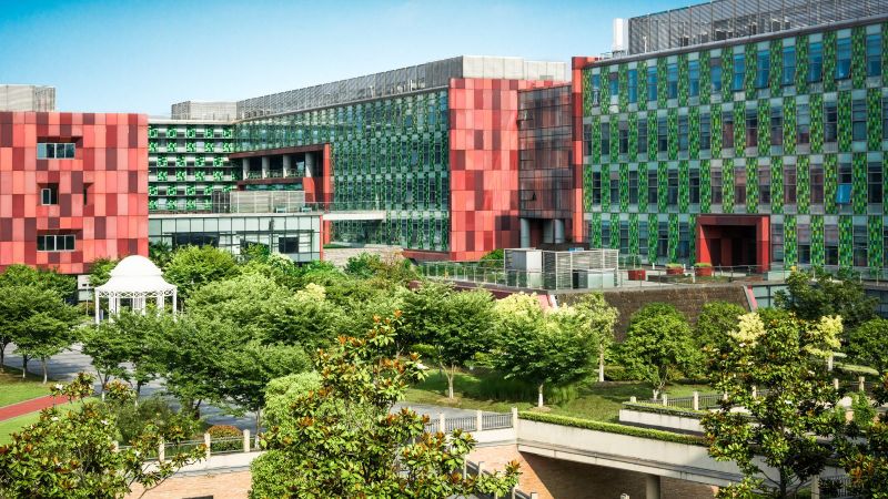 Von Bürogebäuden eingerahmte Grünfläche mit Bäumen, Wegen und einem Pavillon. Im Hintergrund ist blauer Himmel zu sehen.