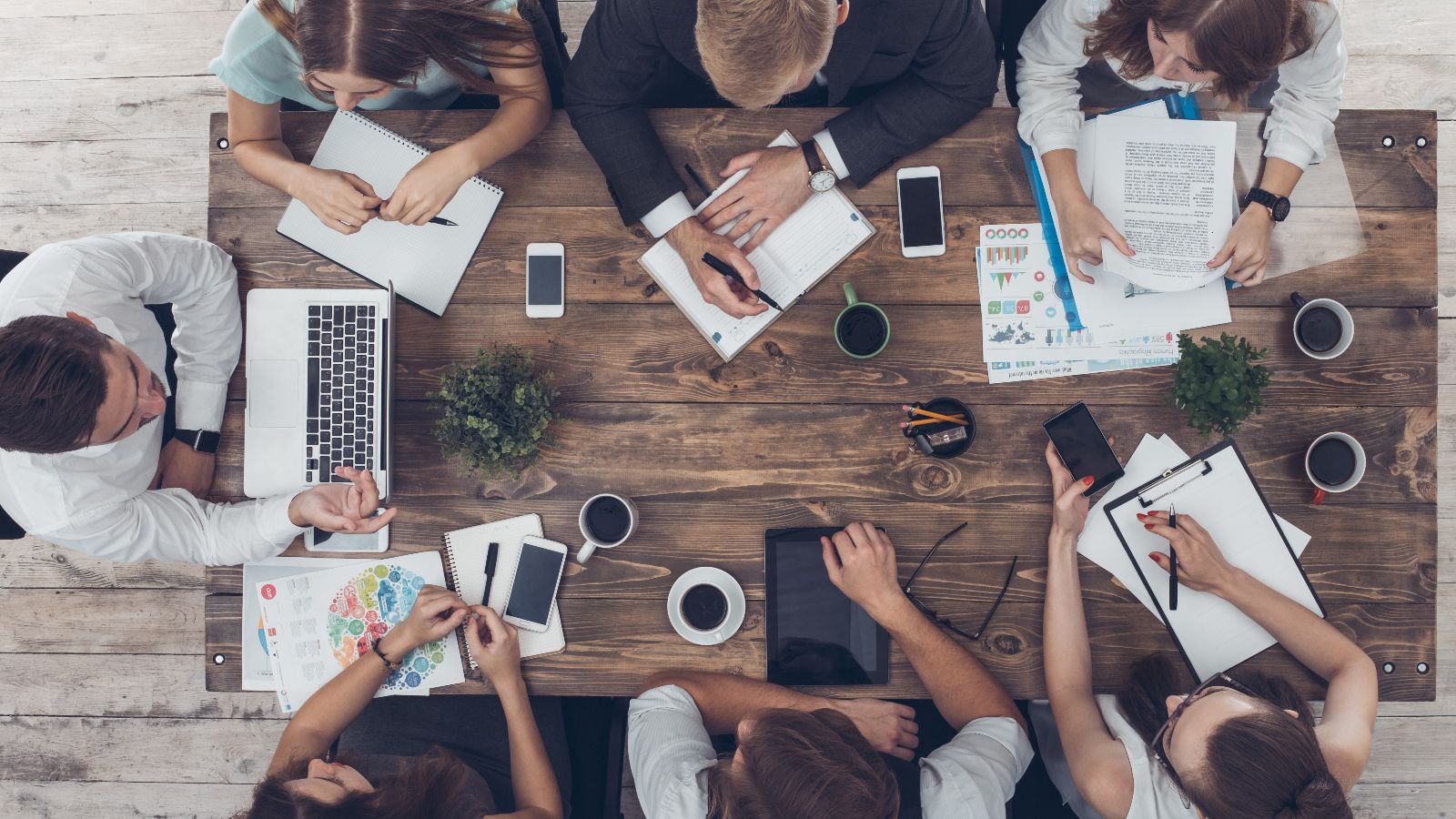 Draufsicht auf einen Tisch mit Unterlagen, Laptops, Tablets, Smartphones und Kaffeetassen. Um den Tisch sitzen sieben Personen und diskutieren.