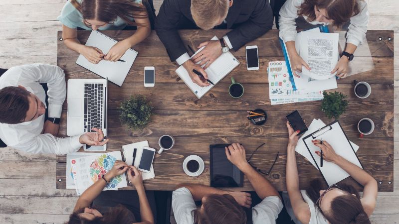 Draufsicht auf einen Tisch mit Unterlagen, Laptops, Tablets, Smartphones und Kaffeetassen. Um den Tisch sitzen sieben Personen und diskutieren.