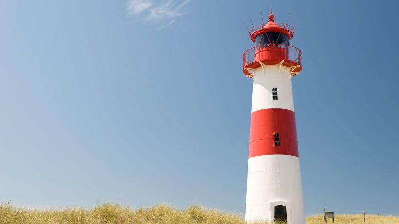 Ein rot-weißer Leuchtturm steht in Dünen vor blauem Himmel.