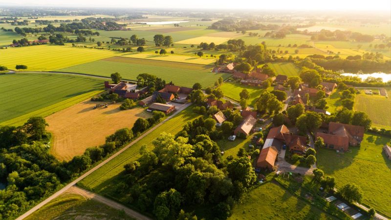 Eine weiträumiger Bauernhof steht inmitten einiger Baumgruppen. Um das Gehöft erstrecken landwirtschaftrliche Flächen über flaches Land bis zum Horizont.