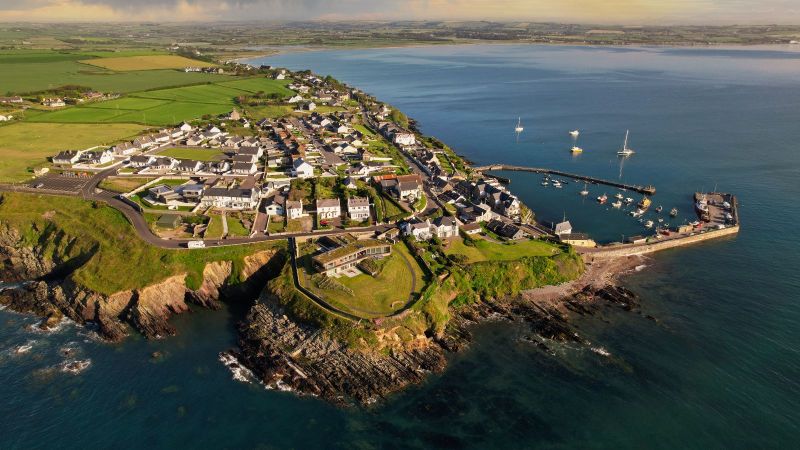 Blick aus der Vogelperspektive auf eine kleine Küstengemeinde mit Hafen. Die Küste ist felsig und in Hintergrund sind grüne Wiesen und Felder zu sehen.