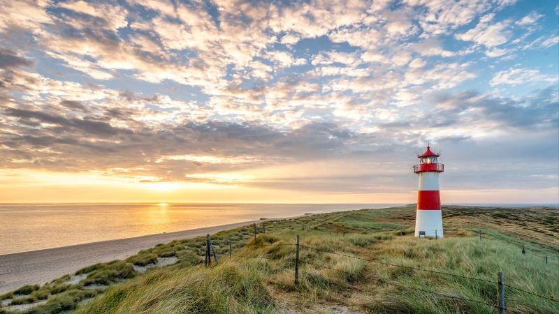 Ein rot-weißer Leuchtturm steht in den Dünen vor einem Strand. Im Hintergrund ist das Meer und der Abendhimmel mit Sonnenuntergang zu sehen.