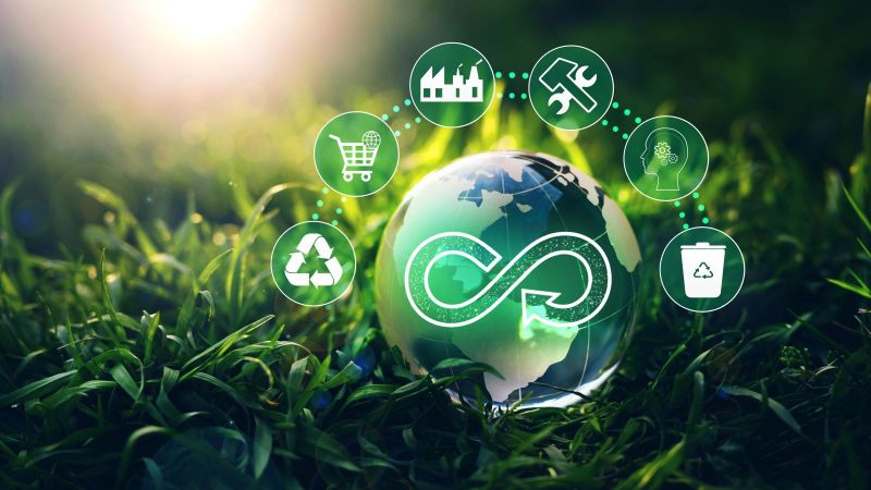 Eine Erdkugel aus Glas liegt im Gras. Auf ihr ist ein Unendlichkeitszeichen, über ihr schweben Symbole zum Thema Kreislaufwirtschaft.