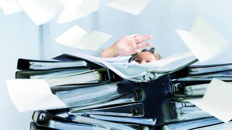 Eine Person ist gerade noch hinter Stapeln von Aktenordnern und fliegenden Zetteln zu sehen.