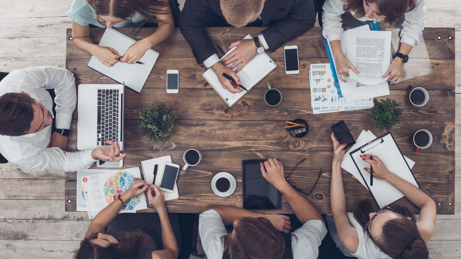 Draufsicht auf einen Tisch mit Unterlagen, Laptops, Tablets, Smartphones und Kaffeetassen. Um den Tisch sitzen sieben Personen und diskutieren.