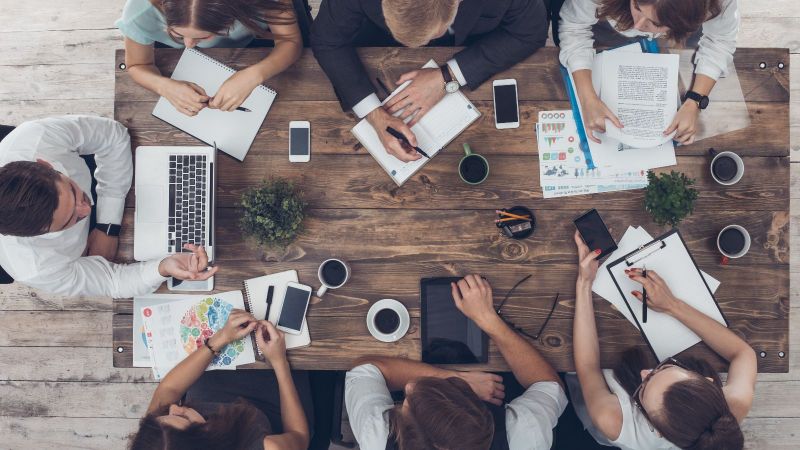 Draufsicht auf einen Tisch mit Unterlagen, Laptops, Tablets, Smartphones und Kaffeetassen. Um den Tisch sitzen sieben Personen und diskutieren.