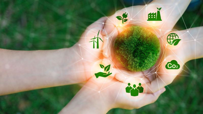 Zwei Paar Hände liegen aufeinander. Darin liegt abstrakt ein aus Moos bestehender Kreis um den Symbole wie Windräder und CO2 angeordnet sind