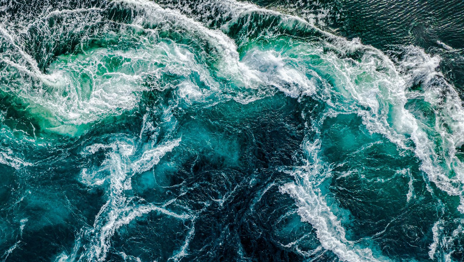 Aus der Vogelperspektive fällt der Blick auf das dunkelblaue Wasser eines aufgewühlten Meeres. Die Gischt bildet ein weißes Muster auf der Wasseroberfläche.