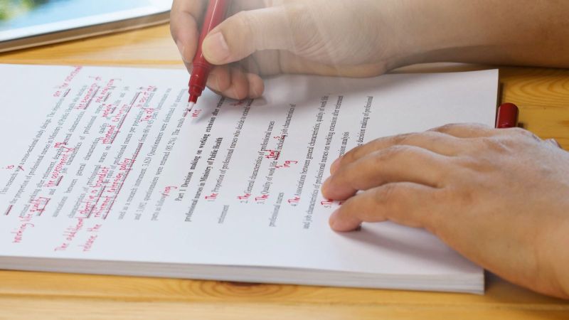 Auf einem Tisch liegt ein Dokument. Darüber sind zwei Hände. Eine Hand hält einen Rotstift und macht Korrekturen, die andere hält die Stiftkappe. Neben dem Dokument liegt ein Handy.