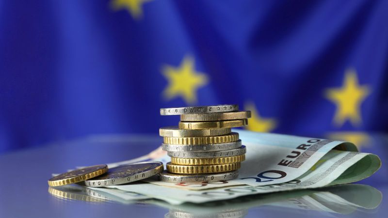 Ein Euro-Münzenstapel liegt auf Euro-Geldscheinen vor einer EU-Flagge