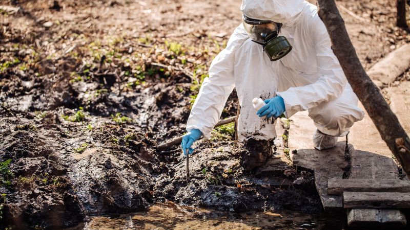 Wissenschaftlerin in Sicherheitsanzug sammelt Bodenprobe in einem Probengefäß. Matschige, rurale Umgebung.