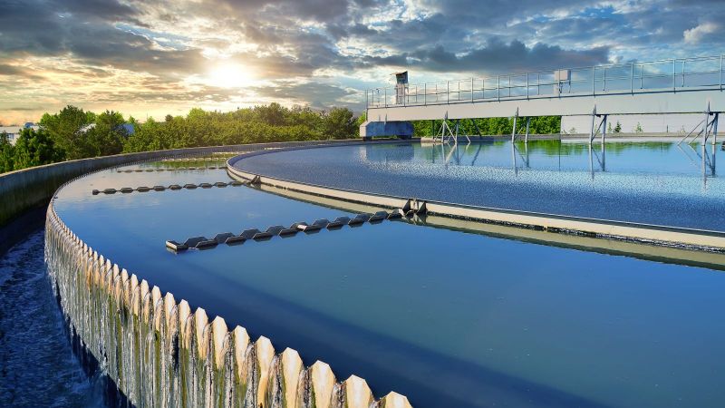 Das Licht der tief stehenden Sonne wird durch die glatte Wasseroberfläche in einer Kläranlage reflektiert. Im Hintergrund sind mehrere Baumkronen zu erkennen.