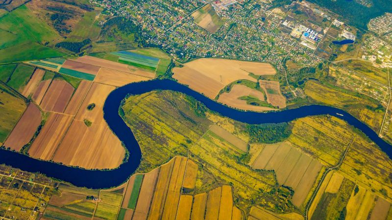 Vogelperspektive auf einen Fluss, welcher durch landwirtschaftlich genutzte Flächen mäandert. Am oberen Rand ist ein Siedlungsgebiet zu sehen