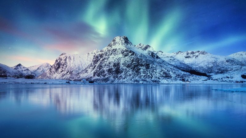 Vor einem stillen Wasser steht ein schneebedeckter Berg. Dahinter sind Nordlichter zu sehen. 