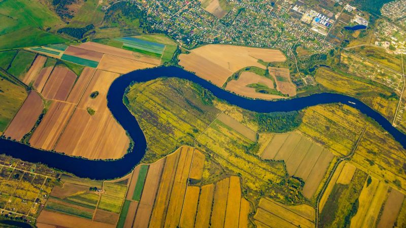 Vogelperspektive auf einen Fluss, welcher durch landwirtschaftlich genutzte Flächen mäandert. Am oberen Rand ist ein Siedlungsgebiet zu sehen.  