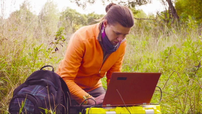 Eine Frau in Trekking-Garderobe hockt im hohen Grass. Neben ihr liegt befindet sich ein Rucksack. Vor ihr steht ein aufgeklappter Laptop auf einer großen Plastikkiste.