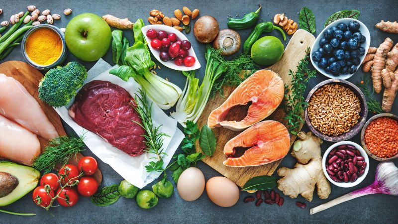 Eine dunkle Arbeitsfläche ist flächendeckend mit diversen frischen Lebensmitteln bedeckt.  Die Lebensmittelauswahl erstreckt sich von Obst und Gemüse neben Fleisch, Fisch und Eiern.