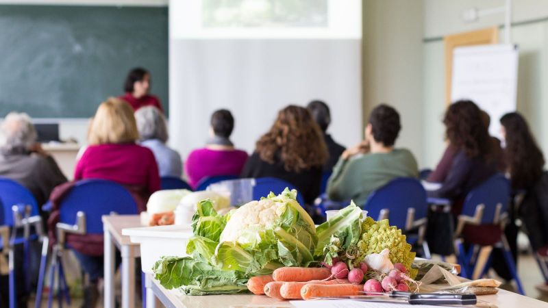 Fokus auf frischem Gemüse auf einem Tisch. Im Hintergrund ist ein sitzendes Publikum bei einem Vortrag in einem Klassenzimmer zu sehen.