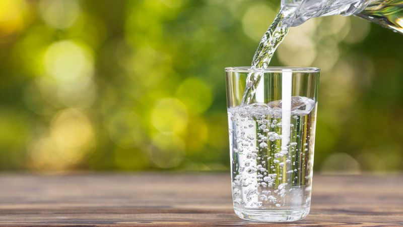 Aus einer Glaskaraffe wird Wasser in ein durchsichtiges Glas gegossen, das auf einem Holztisch steht.