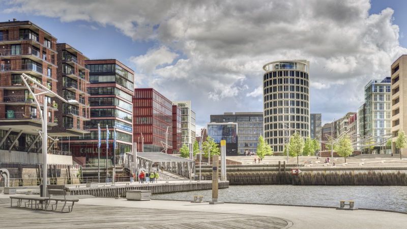 Der Blick fällt auf eine moderne Stadt über ein Wasser mit Bänken am gestalteten Ufer.