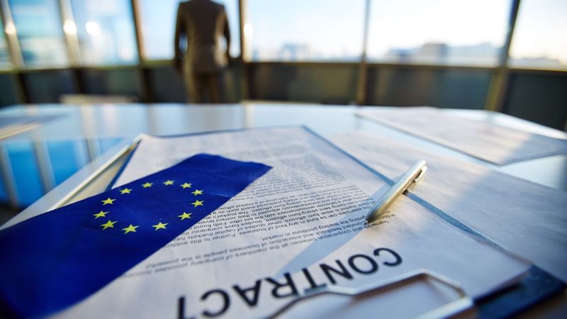 Ein Vertrag liegt auf einem Tisch. Auf dem Vertrag liegen ein Stift und eine EU-Flagge. Im Hintergrund steht eine aus dem Fenster blickende Person.