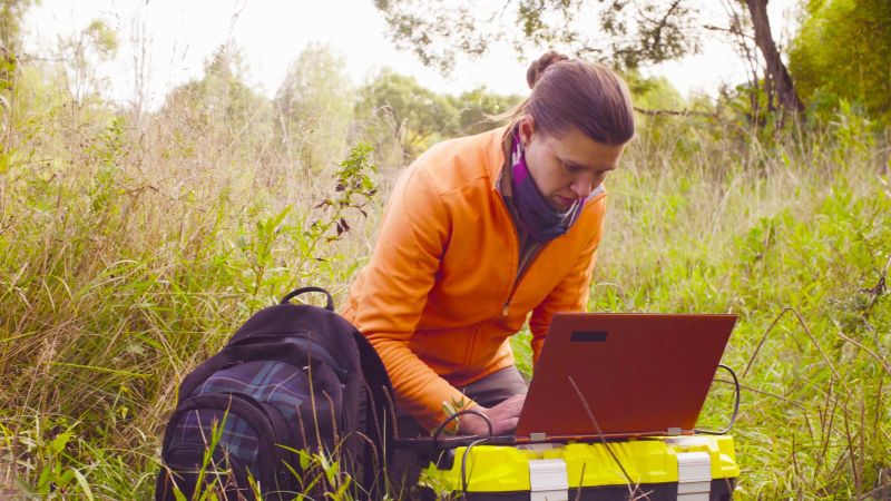 Eine Frau in Trekking-Garderobe hockt im hohen Gras. Neben ihr liegt befindet sich ein Rucksack. Vor ihr steht ein aufgeklappter Laptop auf einer großen Plastikkiste.