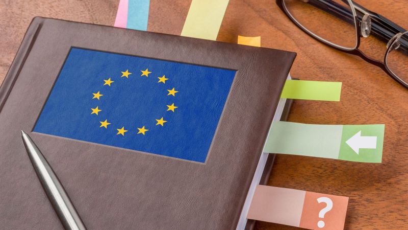 Ein Notizbuch mit einer EU-Flagge als Logo und eine schwarze Lesebrille liegen auf einem Tisch. Zwischen den Seiten im Notizbuch sind bunte Post-its eingeklebt. Auf dem Notizbuch liegt ein Stift.