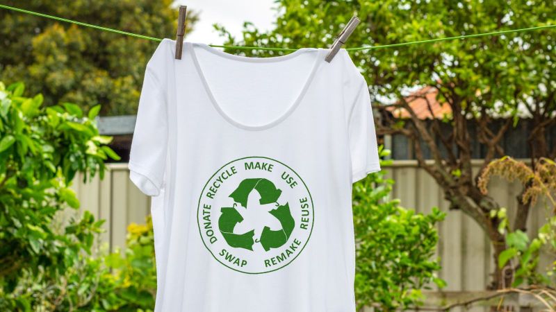 Ein weißes T-Shirt mit einer grünen Abbildung eines Recyclingkreislaufs hängt auf einer Leine in einem grünen Garten