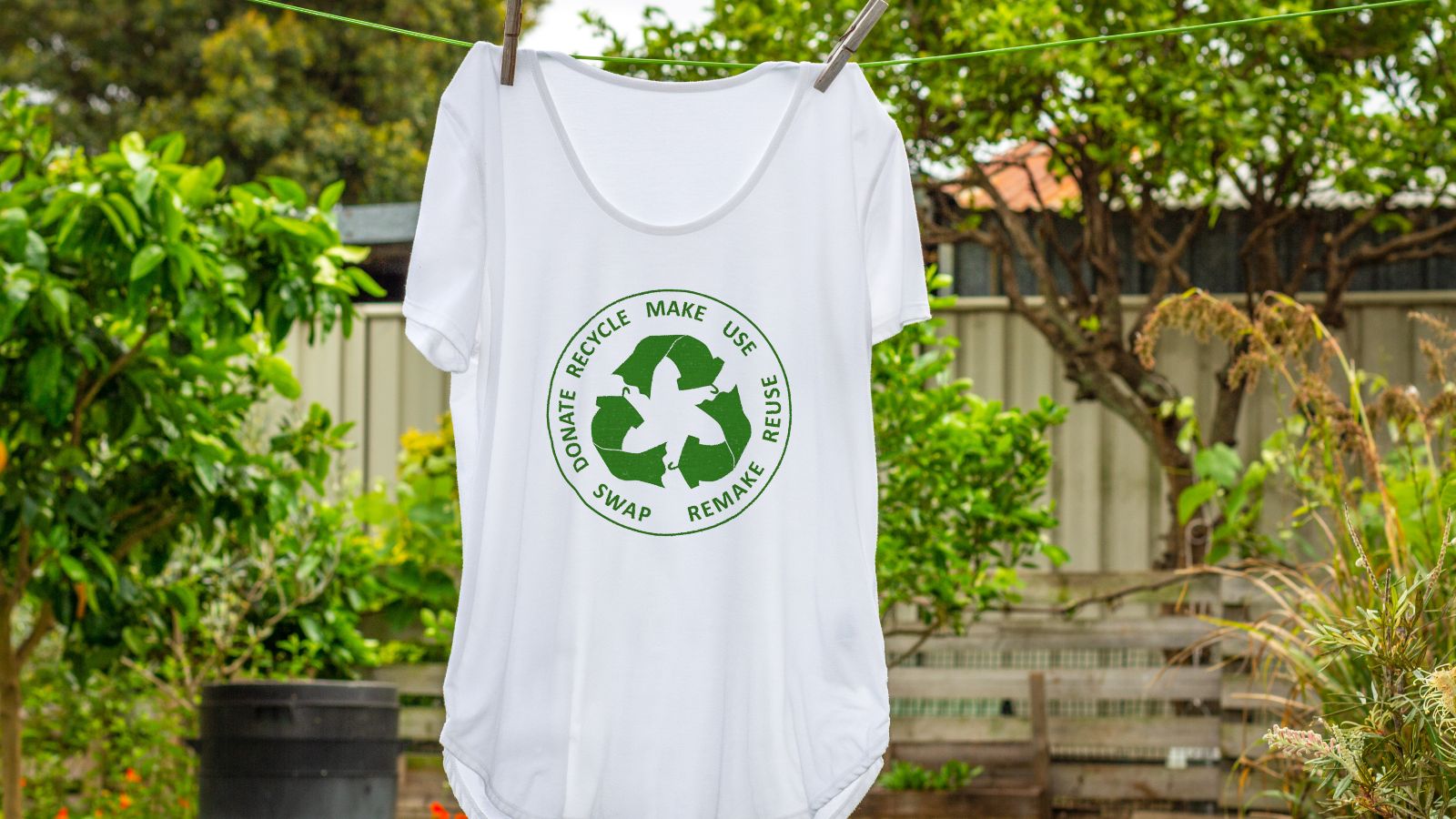 Ein weißes T-Shirt mit einer grünen Abbildung eines Recyclingkreislaufs hängt auf einer Leine in einem grünen Garten.