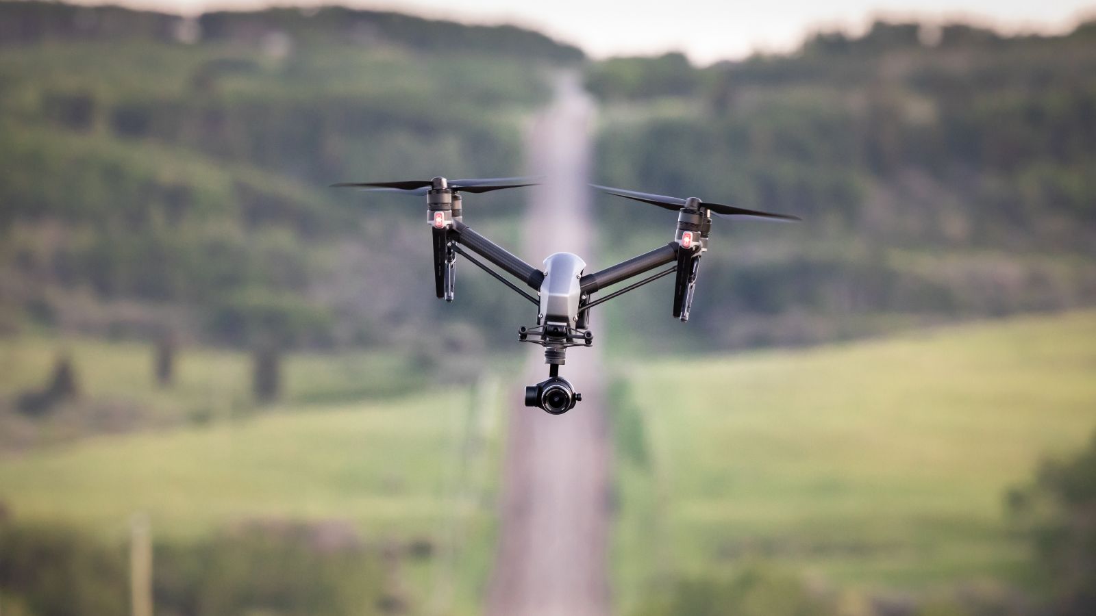 Drone fliegt über eine Landschaft. Im unscharfen Hintergrund ist eine Landstraße erkennbar, welche bergauf führt und von Wald und Wiesen umgeben ist.