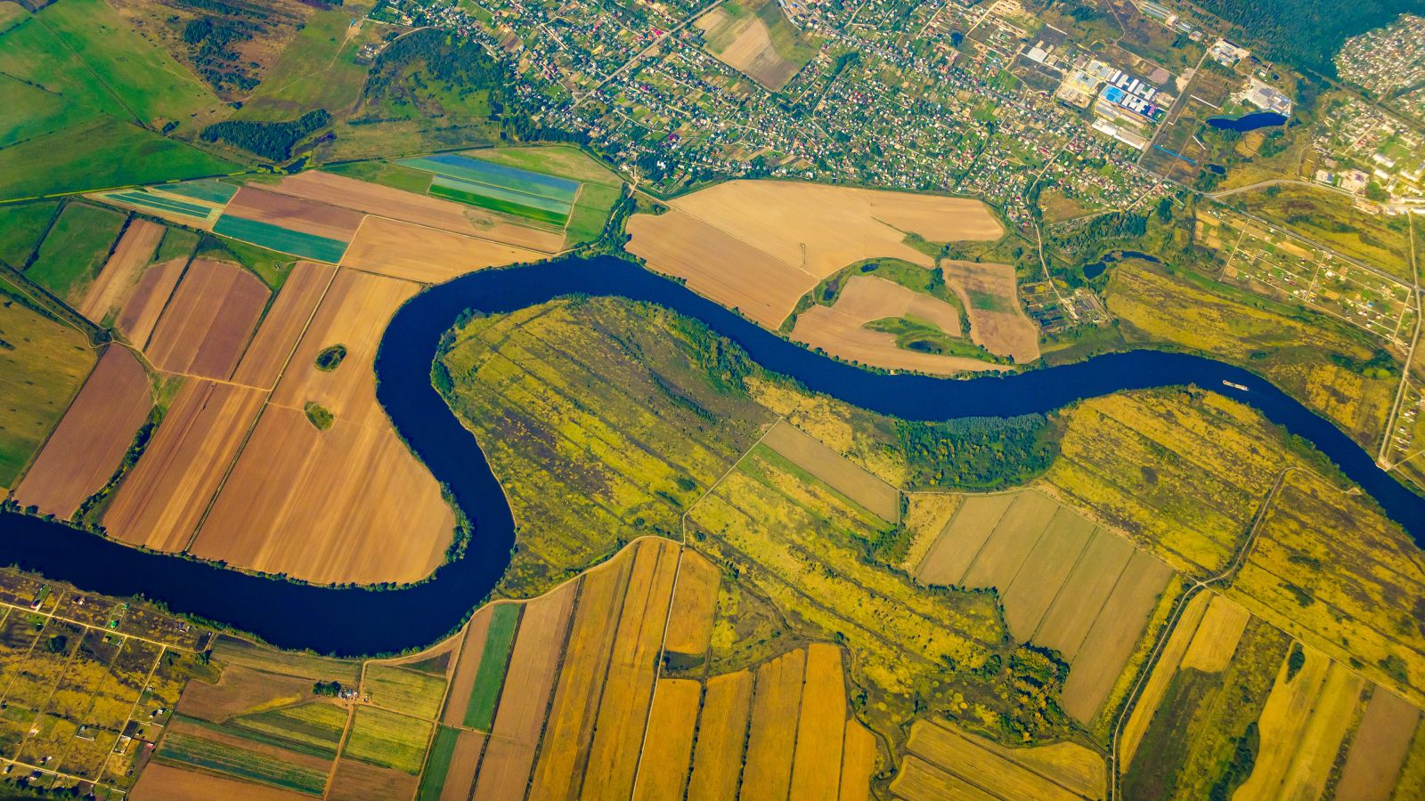 Vogelperspektive auf einen Fluss, welcher durch landwirtschaftlich genutzte Flächen mäandert. Am oberen Rand ist ein Siedlungsgebiet zu sehen.