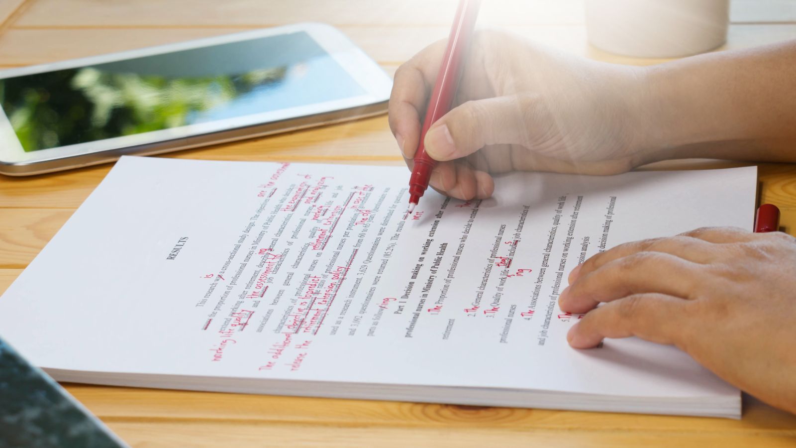 Auf einem Tisch liegt ein Dokument. Darüber sind zwei Hände. Eine Hand hält einen Rotstift und macht Korrekturen, die andere hält die Stiftkappe. Neben dem Dokument liegt ein Handy.